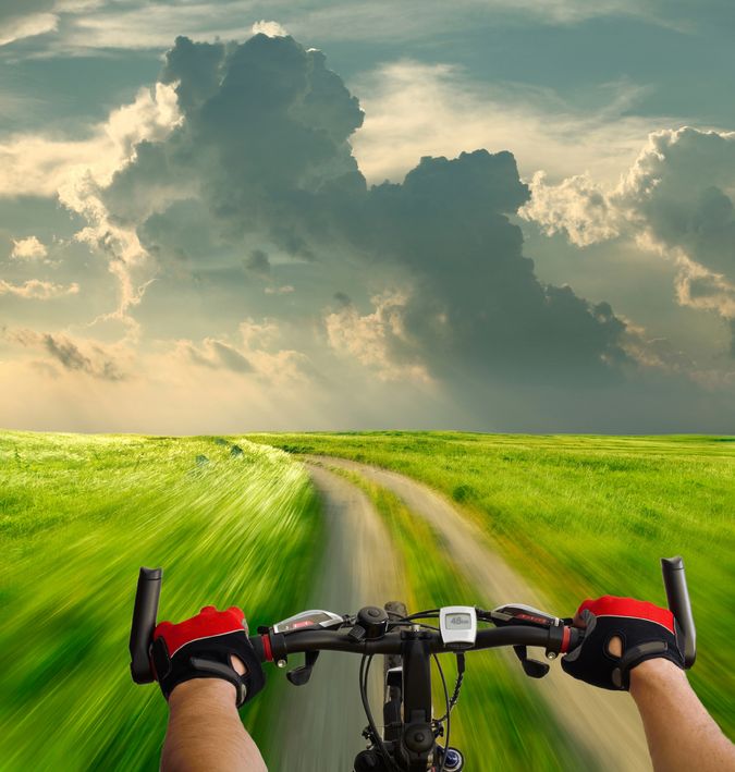 Cyclist's perspective on a scenic dirt path, speeding through lush green fields under a dramatic sky, capturing the thrill of adventure and exploration.