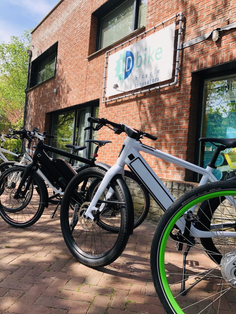 Row of IDbike electric bicycles parked outside IDbike headquarters with company sign on brick building.
