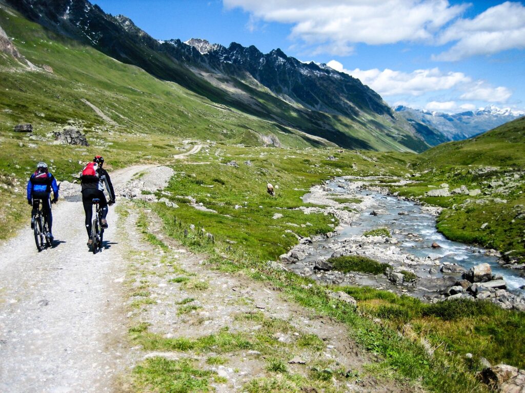 Two cyclists ride on a mountain trail alongside a river with alpine peaks in the background.