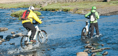 Two riders crossing a stream on electric bikes, showcasing the durability and performance of IDbike e-bikes in outdoor environments